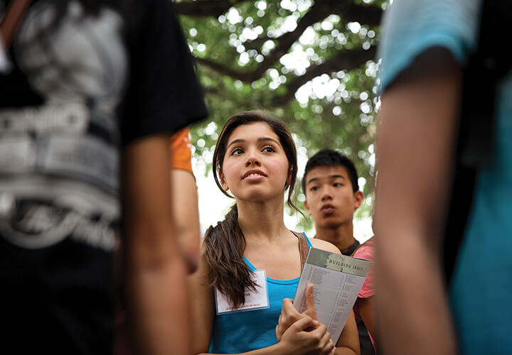 Focus on a female student holding a booklet. Looking excited in the west mall under trees.