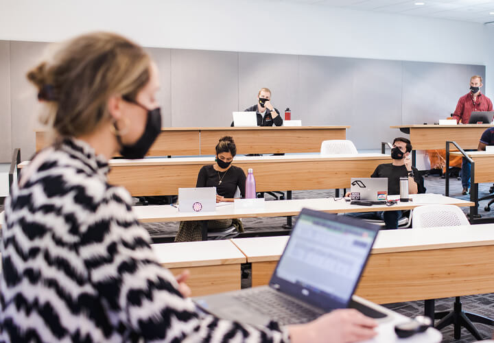 Masked instructor works on laptop in front of class