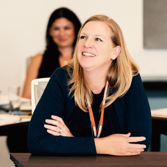 Women sits in class and looks upfront smiling.